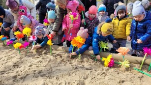 Dzieci pochylają się nad piaskownicą i wkładają w piasek kwiaty z papieru.