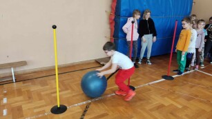 Na sali gimnastycznej chłopiec w czerwonych spodniach turla dużą niebieską piłkę pomiędzy pachołkami.