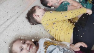 Dzieci leżą na plecach na podłodze obok siebie. Śmieją się. Na nimi rozciągnięta jest przezroczysta folia, na której widac kropelki wody.