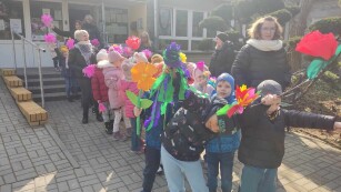 Dzieci z grupy Pszczółek stoją usmiechnięte przed przedszkolem. W rękach trzymają kolorowe kwiaty z papieru.