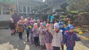 Uśmiechniete dzieci z grupy Motylków stoją przed przedszkolem i machają kolorowymi kwiatkami.