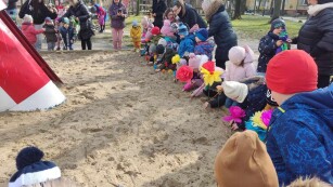 Usmiechnięte dzieci stoją wokół piaskownicy. Wtykają w piasek kolorowe kwiaty z papieru.