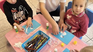 chłopiec w czarnej koszulce i dziewczynka w różpwej bluzce siedzą przy stoliku i kleją łabędzia z papieru, pomaga im pani w białej koszulce