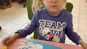 dziewczynka w okularkach  siedzi przy plastikowym stoliku i pokazuje swojego łabędzia z papieru