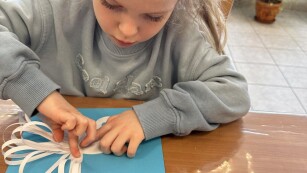 dziewczynka w szarej bluzie siedzi przy stoliku i przykleja białe paski papieru do łabędzia