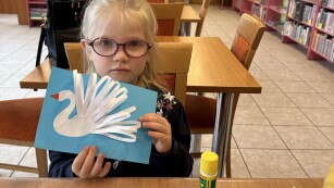 dziewczynka w okularach siedzi przy stoliku a w rękach trzyma białego łabędzia z papieru