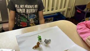 chłopiec w szarej koszulce siedzi przy stoliku i pokazuje zwierzątka z plasteliny