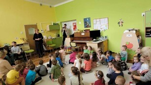 W sali o zielonych ścianach, grupa dzieci siedzi na dywanie i w skupieniu słucha chłopca grającego na pianinie
